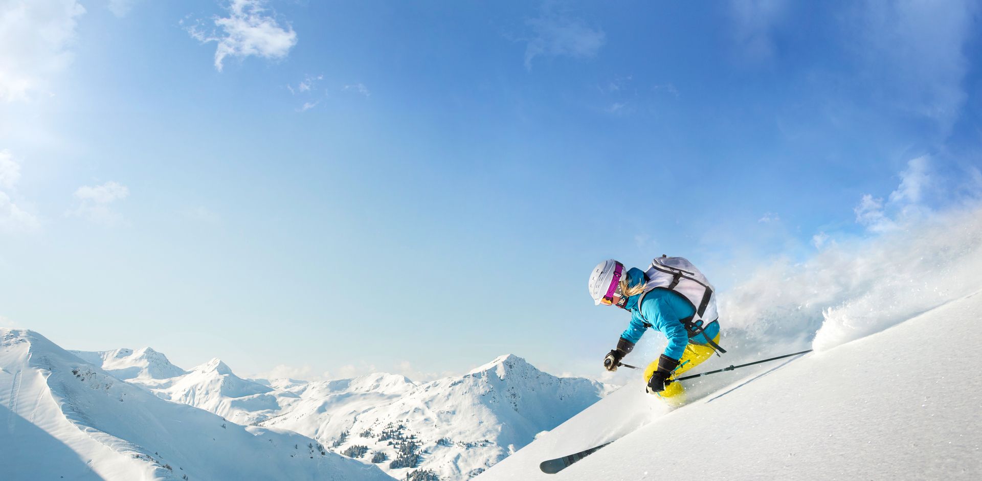 Skifahrer in bunter Kleidung fährt auf verschneiter Piste vor Bergkulisse unter blauem Himmel